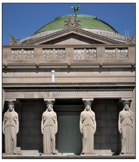 Four stone columns in the shape of women dressed in drapery stand at the front of a stone building with a green, domed roof.