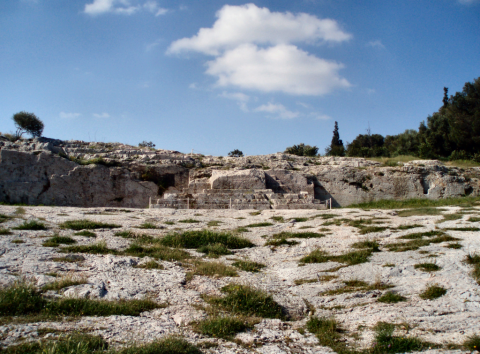 Stone ground with patches of green grass growing through its cracks, with a raised stone platform at one end. The sky is blue with two white clouds.