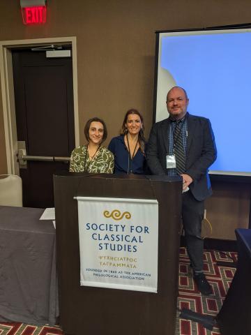 Two women and a man stand smiling behind a podium with a banner that says Society for Classical Studies