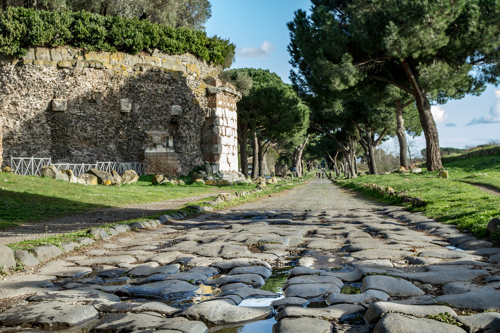 An ancient round mausoleum is on the left, made of brick with bushes growing on top. On the right is a stone road with trees on either side.