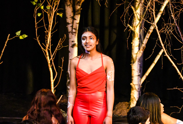 A young woman in a red tank top and pants, her face painted half with white designs, stands in front of a black backdrop with trees.