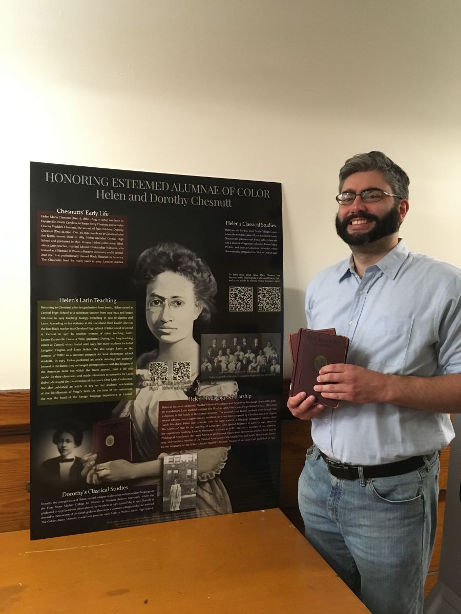 A man holding a book stands next to a poster. The man has dark hair, a dark beard, and glasses and wears a blue short-sleeve shirt, a brown belt, and jeans. The poster is black with a painting of a woman with round metal glasses and assorted text boxes.