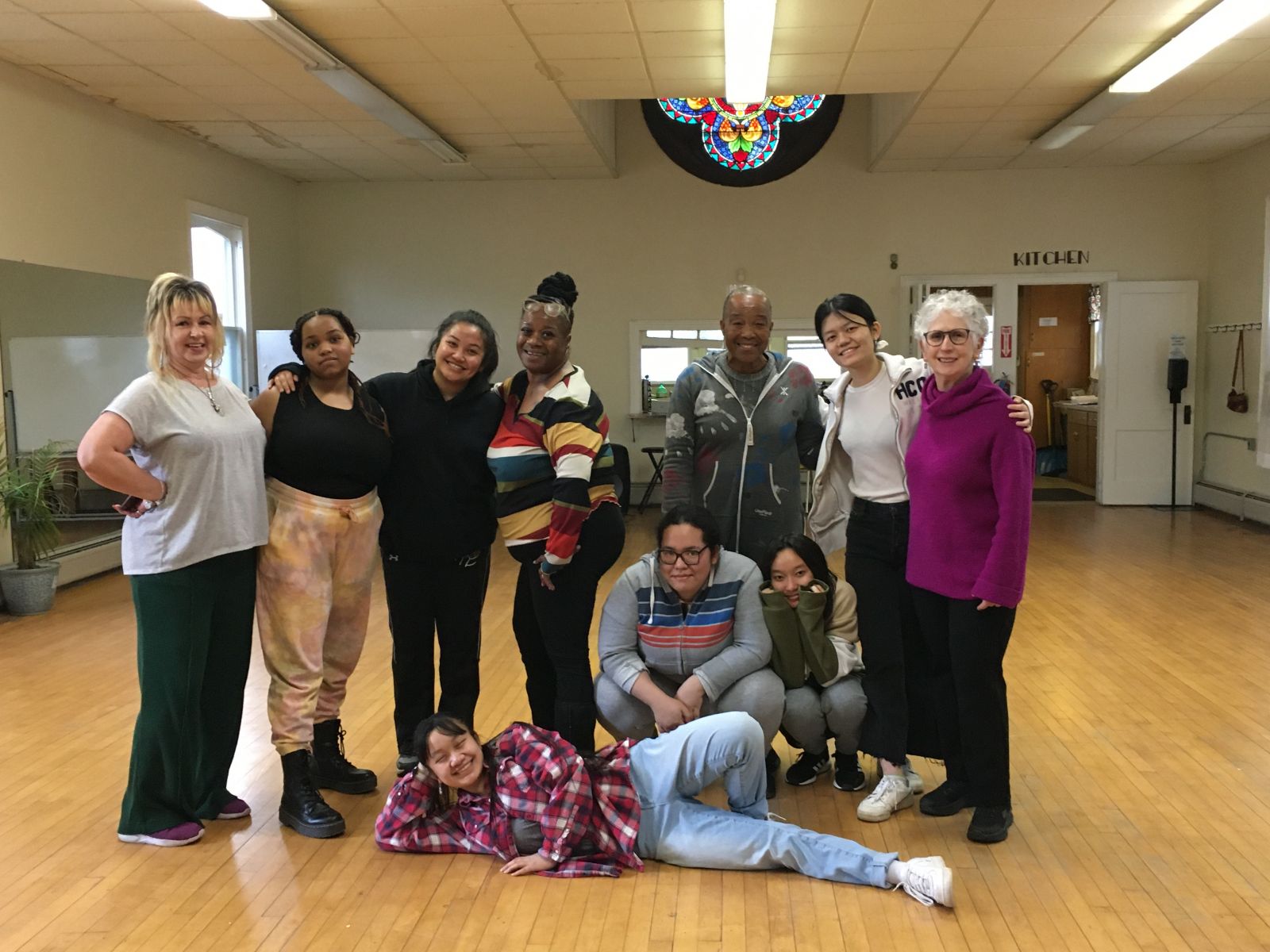 A group of people posing and smiling in a dance studio. Some stand, some squat, and one lies on the ground.