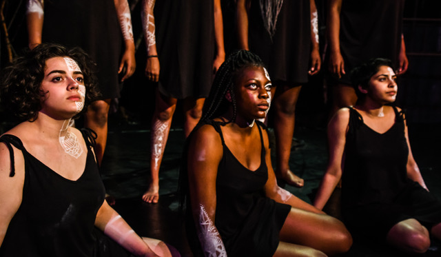 Three young women sit in black sleeveless dresses with half of each woman's face painted with white designs