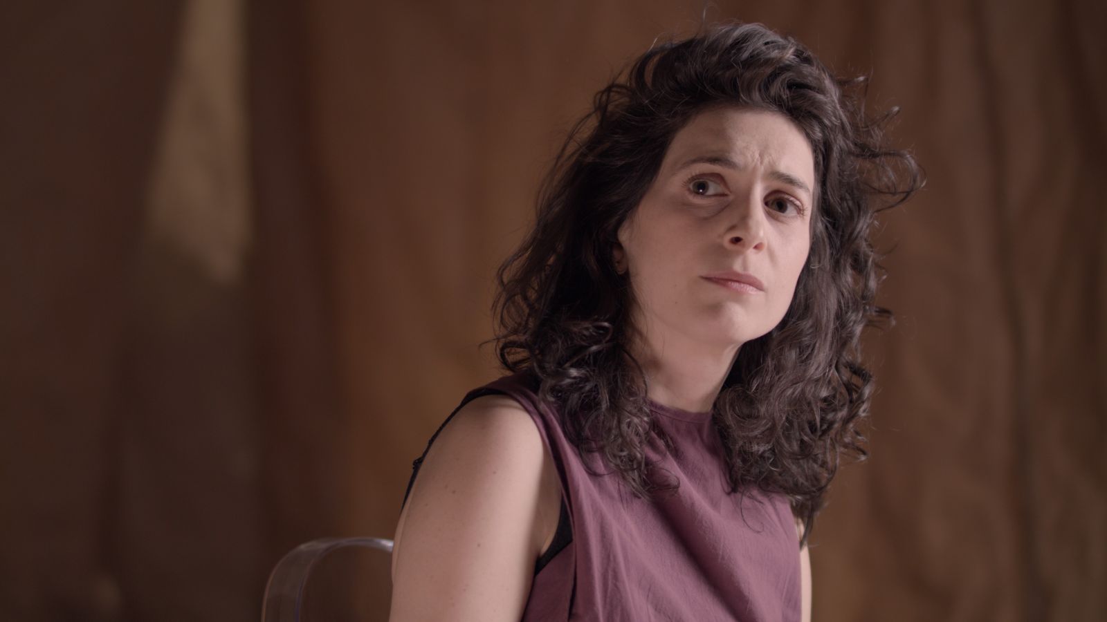 A white woman with dark, curly hair to her shoulders, wearing a mauve sleeveless top, looks sideways.