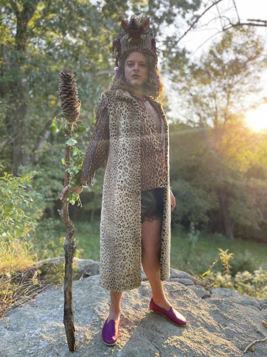 A man with shoulder-length brown hair dresses in a leopard-print blouse, a black miniskirt with lace trim, a long leopard print overcoat, and red shoes. He is carrying a staff and wearing an elaborate headdress.