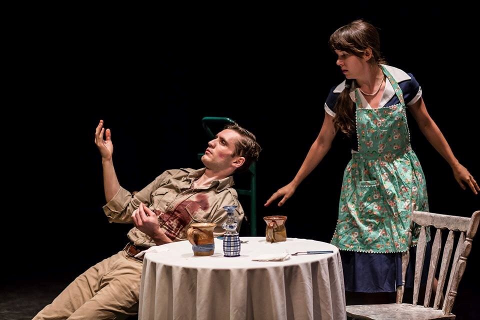 A man in military garb sits at a table. He is raising his hands and has a bloody wound over his left breast. A woman stands behind him wearing an apron.