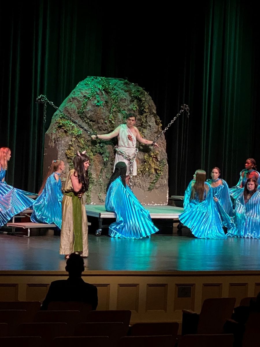 The man stands bound to the rock while a chorus of women in shiny blue pleated dresses circles him.