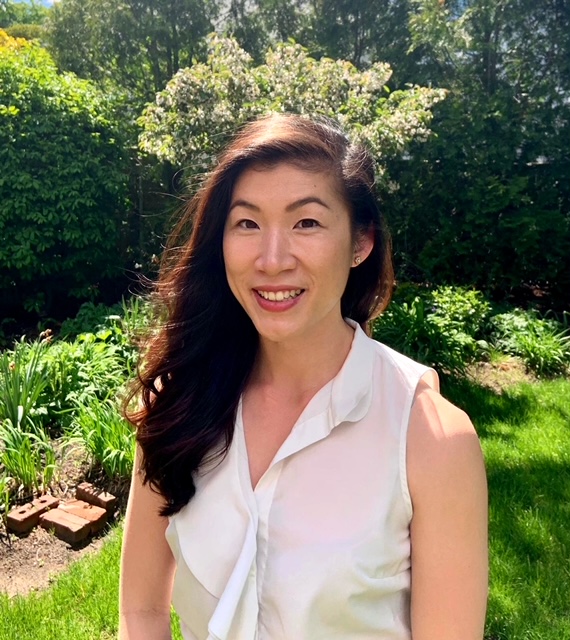Image description: A woman smiling in a white sleeveless blouse stands in front of foliage.
