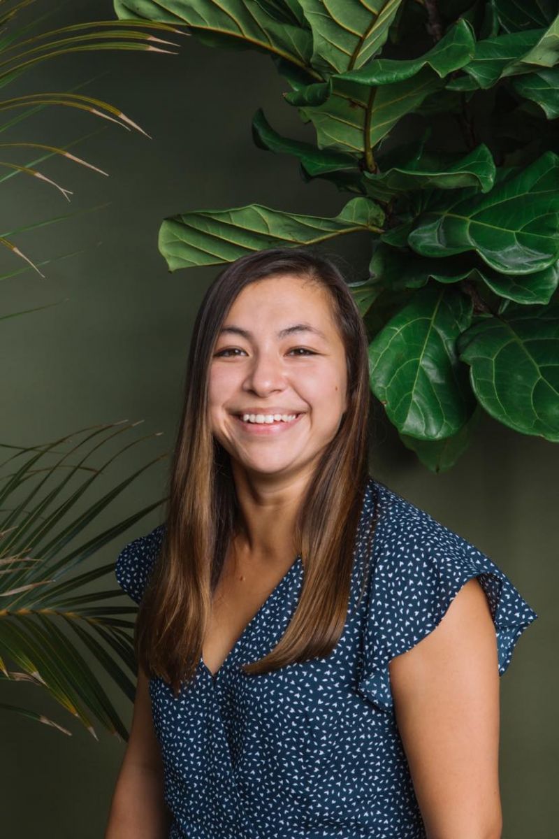 Image description: A woman with straight, shoulder-length hair wearing a blue patterned blouse smiling in front of a palm plant.