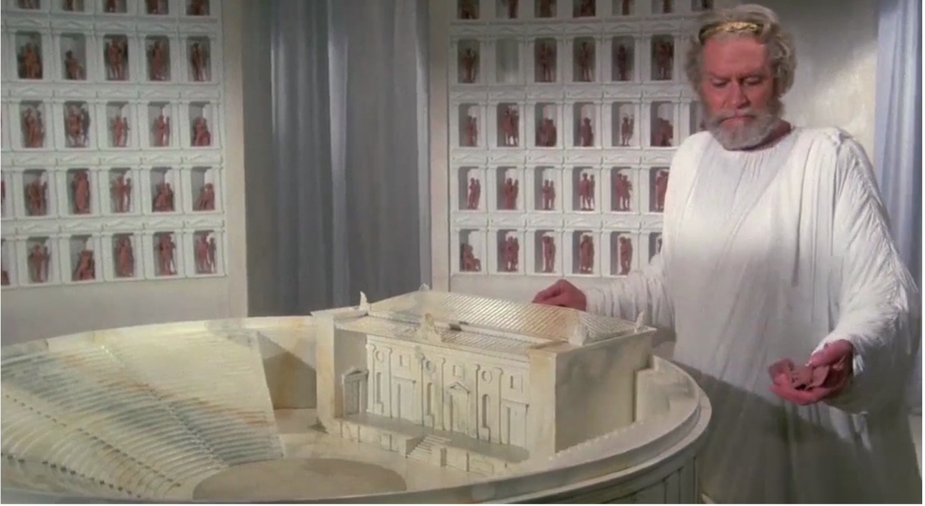 A white man with gray hair and beard wearing a white toga stands over a model of an ancient amphitheater holding people-shaped tokens in his left hand. Behind him are walls with little nooks, within which are more tokens.