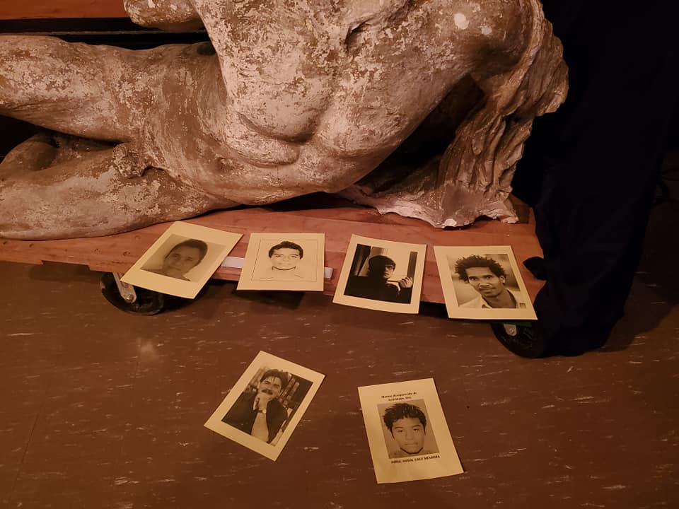 A plaster cast of one of the Parthenon marbles, a male body lying on its left side propped up by its left arm. In front of it are Polaroid photos of people's faces.
