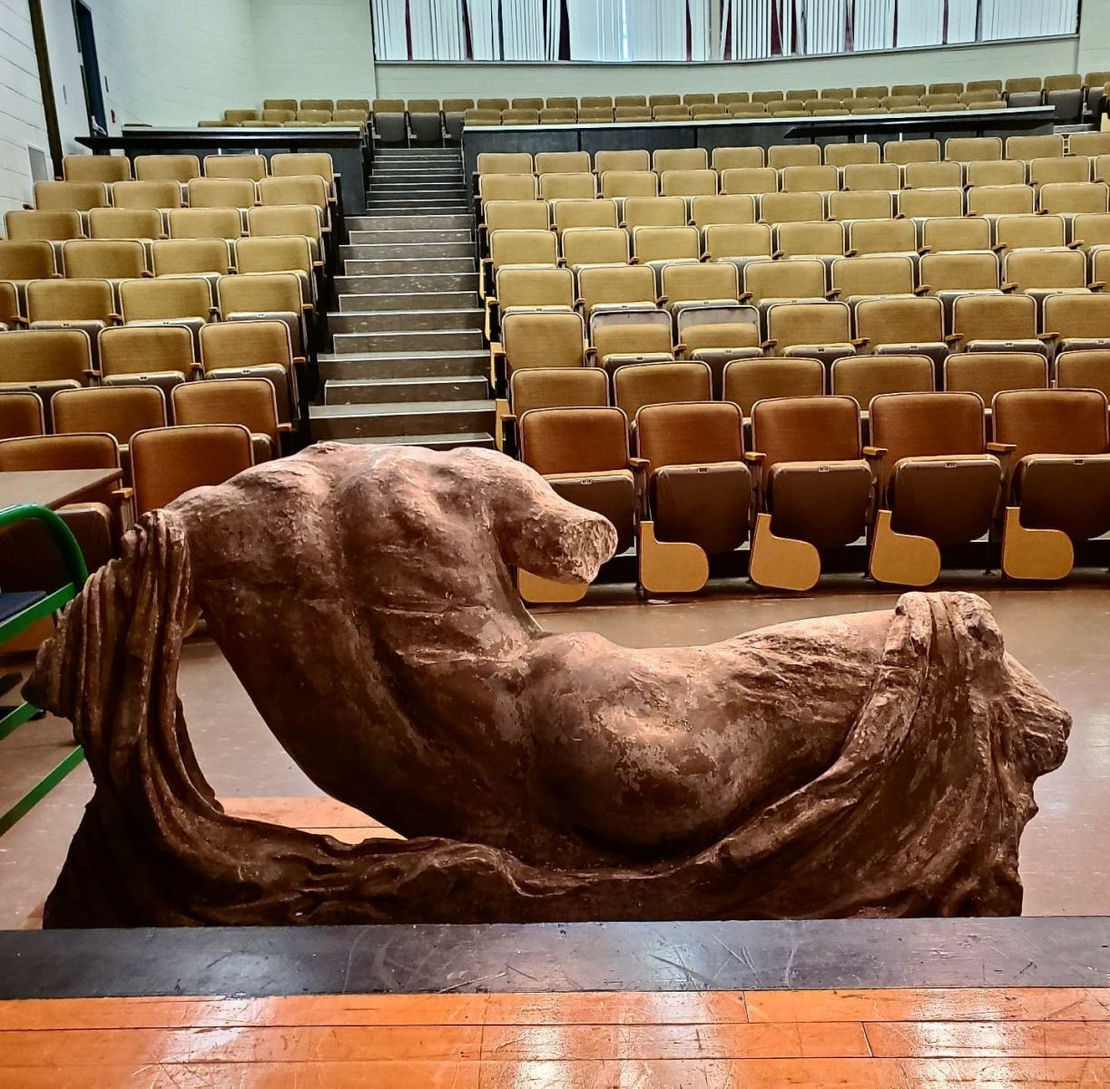 The backside of a plaster cast of one of the Parthenon marbles. A male body lies on its left side,propped up by its left arm which is draped with cloth. The cast sits on stage and faces an empty auditorium of seats.