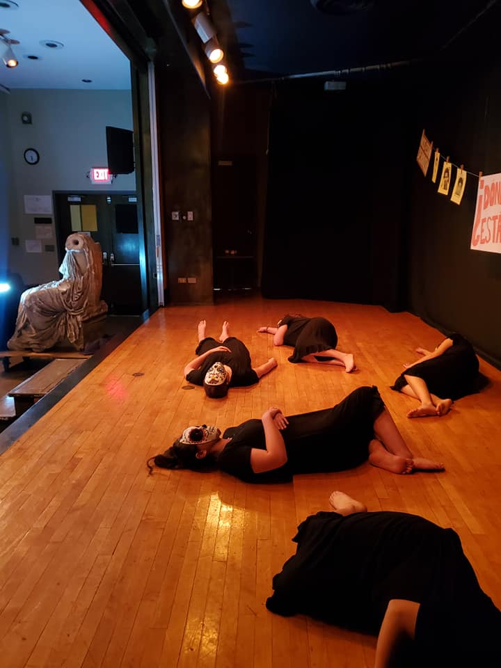 Five women lie onstage wearing black dresses and Dia de los Muertos masks. Their bodies are in different positions.
