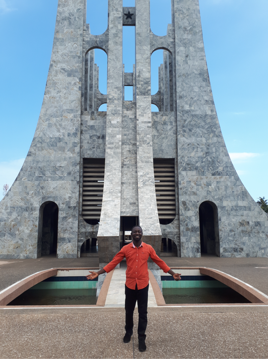 A dark-skinned man with short hair in a red shirt stands in front of a tall stone structure with two small pools on either side. His arms are stretched down and outwards.