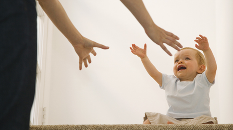 A light-skinned baby with light hair reaches their arms into the air towards a standing adult, whose arms are reaching down to the baby.