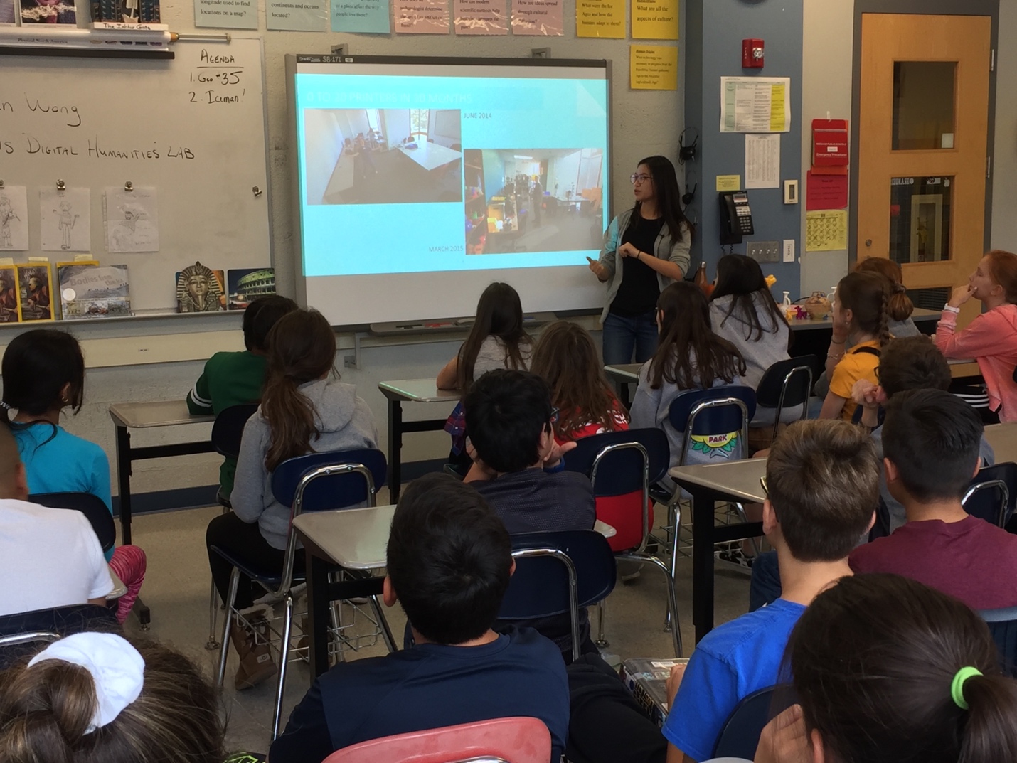 A woman in a black shirt, gray hoodie, and blue jeans with long, dark hair stands in front of a Smartboard in a classroom. Students seated at desks watch her.