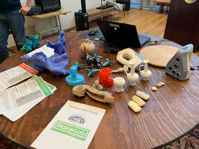A round, brown wooden table with papers and a laptop computer on top. Also on top are 3D printed replicas of ancient artifacts in various colors.
