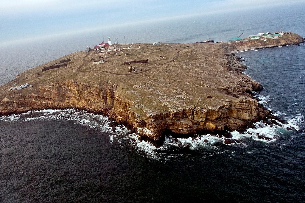 A small island covered in grass with a few buildings sits in the middle of the sea