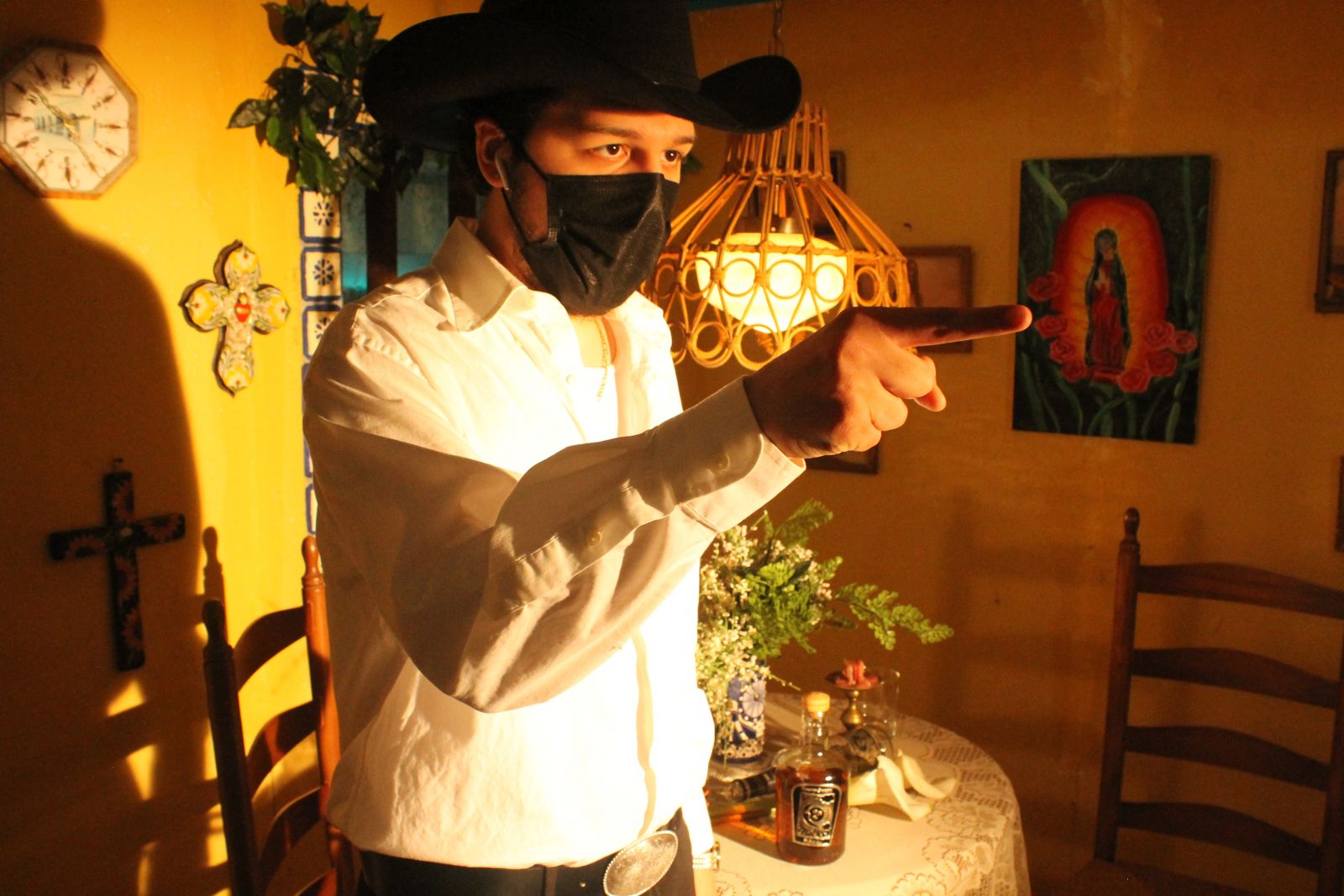 A man wearing a white dress shirt, a black cowboy hat, and a black face mask stands in a room with decorated walls. He points ahead of him.